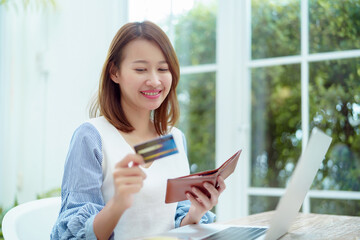 Fototapeta premium A beautiful Asian woman wearing a white shirt with a credit card comes out of her wallet smiling happily in front of her laptop preparing for online shopping.