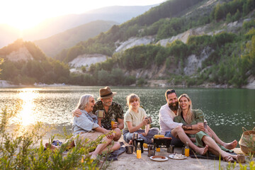 Happy multigeneration family on summer holiday trip, barbecue by lake at sunset.