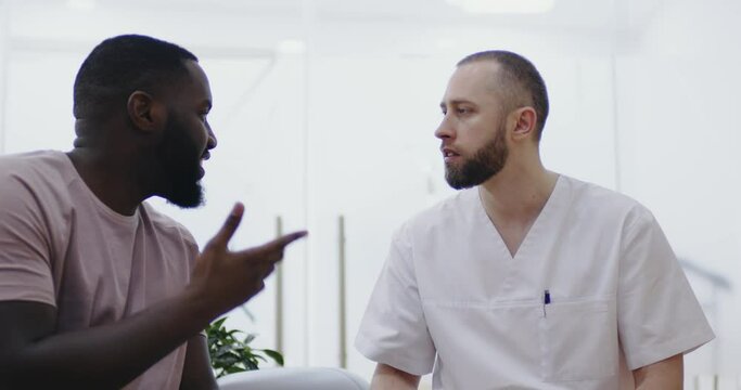 A close-up male medical worker is sitting in the dental clinic and looking at the black man who is showing something on his upper teeth.