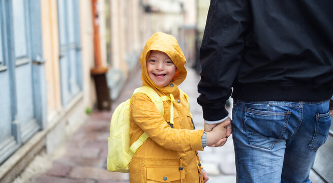 Happy down syndrome boy with unrecognizable father outdoors on a walk in rain, looking at camera.