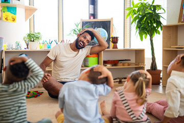 Group of small nursery school children with man teacher sitting on floor indoors in classroom,...