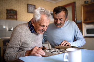 Portrait of man with elderly father sitting at the table indoors at home, looking at photo album.