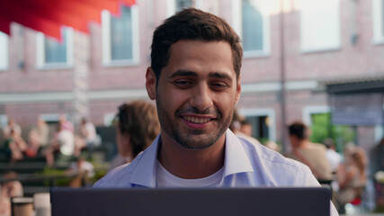 Bokeh shot of indian male having video call on laptop at outdoors terrace of cafe