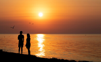 Sunrise on the sea with silhouettes of a young couple and seagulls on the sky background.