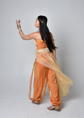 Full length portrait of pretty young asian woman wearing golden Arabian robes like a genie, standing pose  with back to the camera, isolated on studio background.