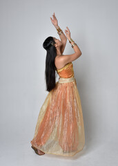 Fototapeta premium Full length portrait of pretty young asian woman wearing golden Arabian robes like a genie, standing pose with back to the camera, isolated on studio background.