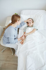 High angle portrait of caring mother by bed of sick child in hospital room, focus on little girl smiling