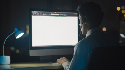 Millennial Asia young call center agent or customer support service executive using computer and microphone headset working technical support in late night office. Telemarketing or sales job concept.