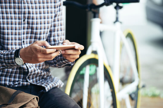 Close-up image of young man sitting next to his bicycle and texting friends or using new popular application on smartphone