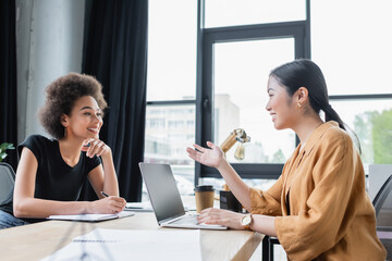 asian businesswoman talking to smiling african american colleague near laptop with blank screen