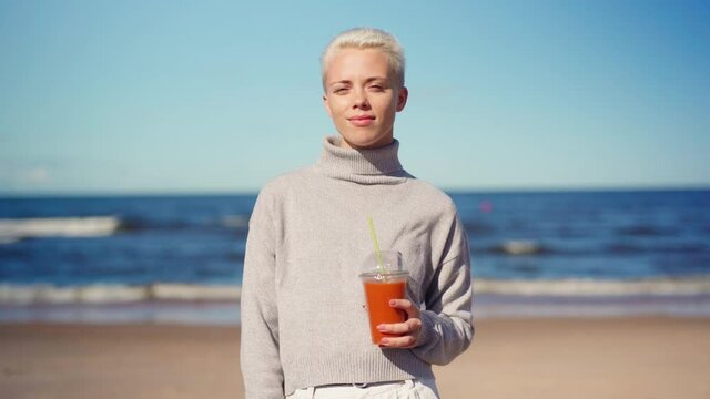 Waist up portrait of young woman with blonde short hair in gray turtleneck sweater standing on beach ooking at camera and smiling with smoothie, juice or cocktail in her hand, sea in background