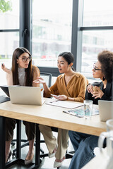 asian businesswoman pointing at laptop while talking to multiethnic colleagues in office