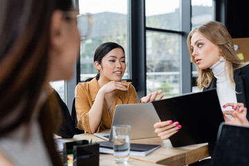 smiling asian businesswoman pointing at laptop near colleague with clipboard, blurred foreground