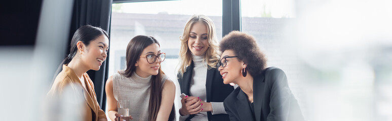 cheerful multicultural businesswomen talking in office on blurred foreground, banner