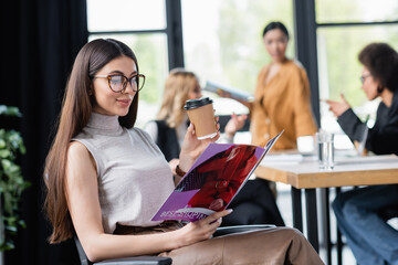 pleased businesswoman with paper cup reading best shopping magazine during coffee break in office