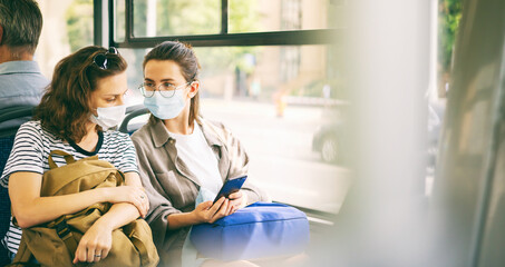 Two young woman friends in protective mask sitting in public bus and reading news in smartphone...