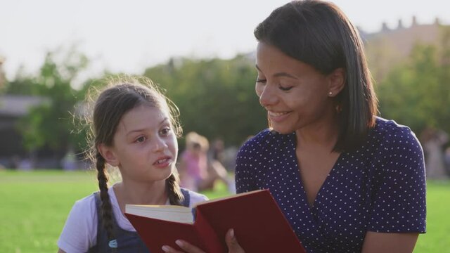 African mother with adopted caucasian daughter sitting on lawn and read book outdoors
