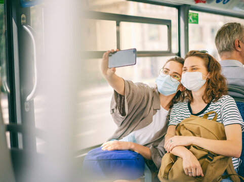Two young woman friends in a protective mask sitting in a public bus and taking a selfie in a smartphone online, traveling during covid