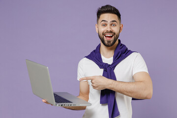 Excited overjoyed beautiful happy smiling young brunet man 20s wears white t-shirt purple shirt hold use work on laptop pc computer pointing on it isolated on pastel violet background studio portrait