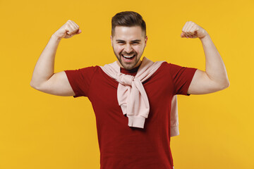 Young overjoyed man 20s in red t-shirt casual clothes showing biceps muscles on hand demonstrating strength power isolated on plain yellow color background studio portrait People lifestyle concept