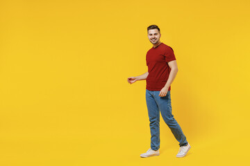 Full length smiling happy caucasian young man 20s wear red t-shirt casual clothes walking going look camera isolated on plain yellow color wall background studio portrait. People lifestyle concept