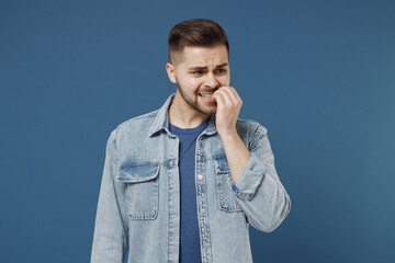 Nervous unnerved aggrieved frowning distempered young brunet man 20s wears denim jacket bite his nails look camera isolated on dark blue background studio portrait. People emotions lifestyle concept.