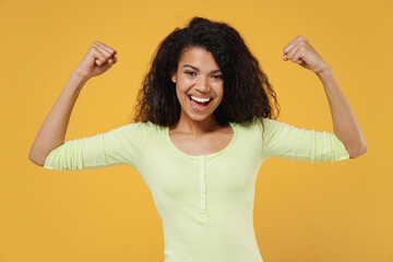 Strong sporty fitness smiling fun african american young brunette woman 20s wears green shirt showing biceps muscles on hand demonstrating strength power isolated on yellow background studio portrait - obrazy, fototapety, plakaty