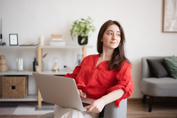Young happy brunette woman in red blouse working on laptop sitting on chair at home in interior