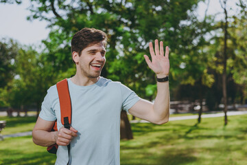 Student young happy friendly man in blue t-shirt backpack waving hand as notice someone walk rest relax in spring green city park go down lawn sunshine outdoor on nature Education high school concept.