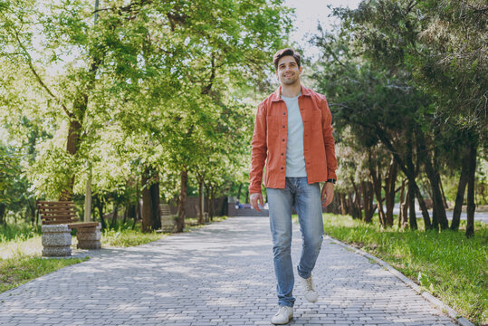 Full length young man 20s wearing orange jacket blue t-shirt walking look camera rest relax in spring green city park go down alley sunshine lawn outdoors on nature. Urban lifestyle leisure concept.