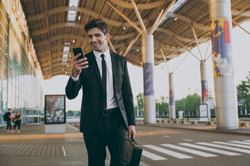 Bottom view fun traveler businessman man in black dinner suit stand outdoorat international airport terminal with attache case hold mobile phone book taxi order hotel Air flight business trip concept