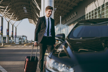 Fototapeta na wymiar Full body bottom view young traveler businessman young man in black dinner suit going walk outside at international airport terminal with suitcase to car booking taxi Air flight business trip concept