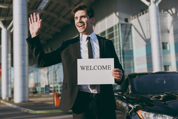 Bottom view young cheerful friendly traveler businessman man 20s in black suit stand outside at international airport terminal hold card sign with welcome title text Air flight business trip concept