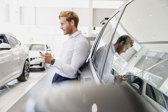 Man side view customer male buyer client in white shirt talk mobile cell phone leaning on car choose auto want buy new automobile in showroom vehicle dealership store motor show indoor Sales concept.