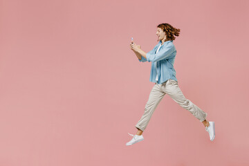 Fototapeta na wymiar Full size side view young excited happy man 20s with long curly hair wear blue shirt white t-shirt using hold in hand mobile cell phone jump high isolated on pastel plain pink color background studio