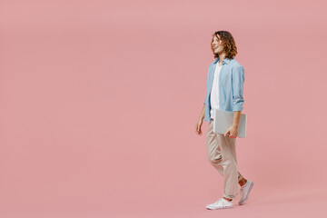 Full length side view young happy man with long curly hair wear blue shirt white t-shirt hold closed laptop pc computer work look aside walk isolated on pastel plain pink color wall background studio.