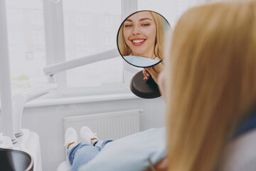 Young smiling beautiful fun happy calm woman 20s reflected in mirror sitting at dental office chair...