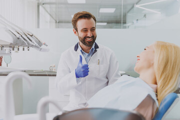 Fototapeta premium Young smiling fun man dentist doctor in white gown show thumb up gesture consulting patient woman sitting at dentist office chair indoor cabinet near stomatologist Healthcare enamel aesthetreatment