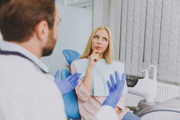 Young man dentist doctor wear white gown talk speak with patient consult minded wistful woman prop up chin sit at dentist office chair indoor cabinet near stomatologist Healthcare enamel treatment