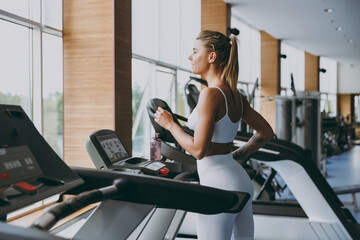 Fototapeta na wymiar Side profile view young skinny strong sporty athletic sportswoman woman in white sportswear warm up training jogging running on a treadmill in gym indoors. Workout sport motivation lifestyle concept.
