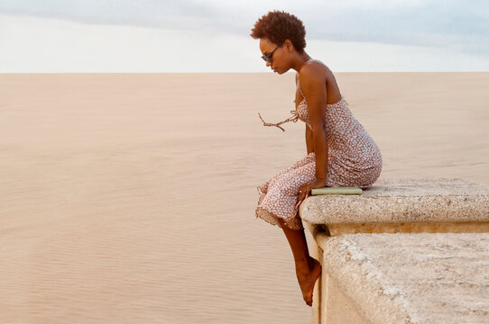 Woman with sunglasses and afro sitting on a wall with a book beside her. Concept of peace and serenity. Latin woman