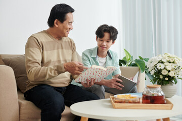 Smiling mature man showing good book to his teenage son when they are drinking tea
