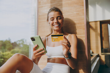 Young happy sportswoman woman in white sportswear earphones listen music use mobile cell phone credit bank card shopping online order delivery book tour pay gym membership sit on windowsill indoor.