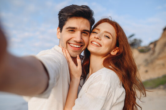Close up smiling happy young couple two family man woman 20s in white clothes hug each other do selfie shot pov on mobile phone at sunrise over sea beach outdoor seaside in summer day sunset evening.