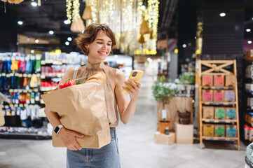 Young woman in casual clothes shopping at supermaket store with craft paper package with groceries using mobile cell phone browsing internet look for recipe inside hypermarket Gastronomy food concept - obrazy, fototapety, plakaty