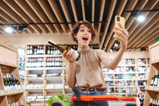 Young excited woman 20s in casual clothes shopping at supermaket store with grocery cart hold wine alcohol hold bottle do selfie shot on mobile phone inside hypermarket Purchasing gastronomy concept