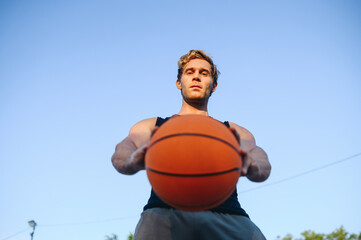 Bottom view young sporty sportsman man in sports clothes look camera training hold in hand ball dribbling play at basketball game playground court on sky background. Outdoor courtyard sport concept