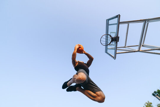Bottom view young strong sportsman man in sports clothes train shooting free throw scoring basket play with ball at basketball game playground court, sky background Outdoor courtyard sport concept
