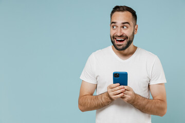 Young smiling happy minded man 20s wear casual basic white t-shirt using mobile cell phone look aside on workspace area mock up isolated on plain pastel light blue color background studio portrait