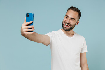 Young smiling fun happy man in white t-shirt doing selfie shot on mobile cell phone post photo on social network isolated on plain pastel light blue color background studio. People lifestyle concept.